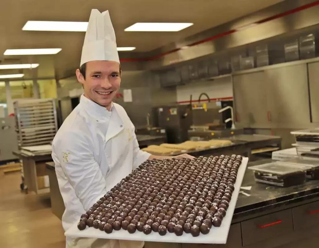 Philipp Zauner stands in the bakery, looks into the camera with a smile and holds up a blob of chocolates.