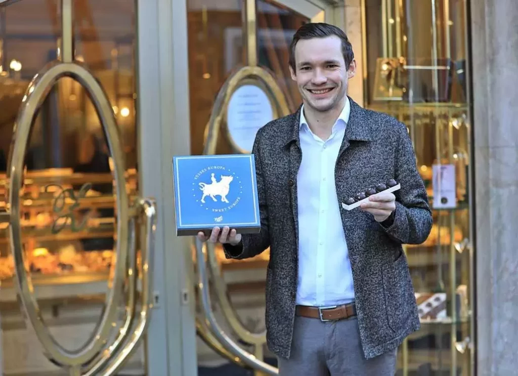 Philipp Zauner in front of the Pfarrgasse entrance, looks into the camera with a smile and holds up a European cake and salt truffle chocolates.