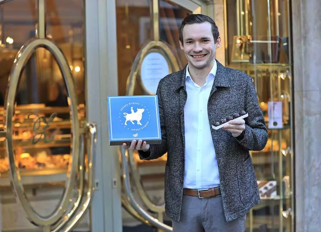 Philipp Zauner in front of the Pfarrgasse entrance, looks into the camera with a smile and holds up a Europa cake and salt truffle chocolates.